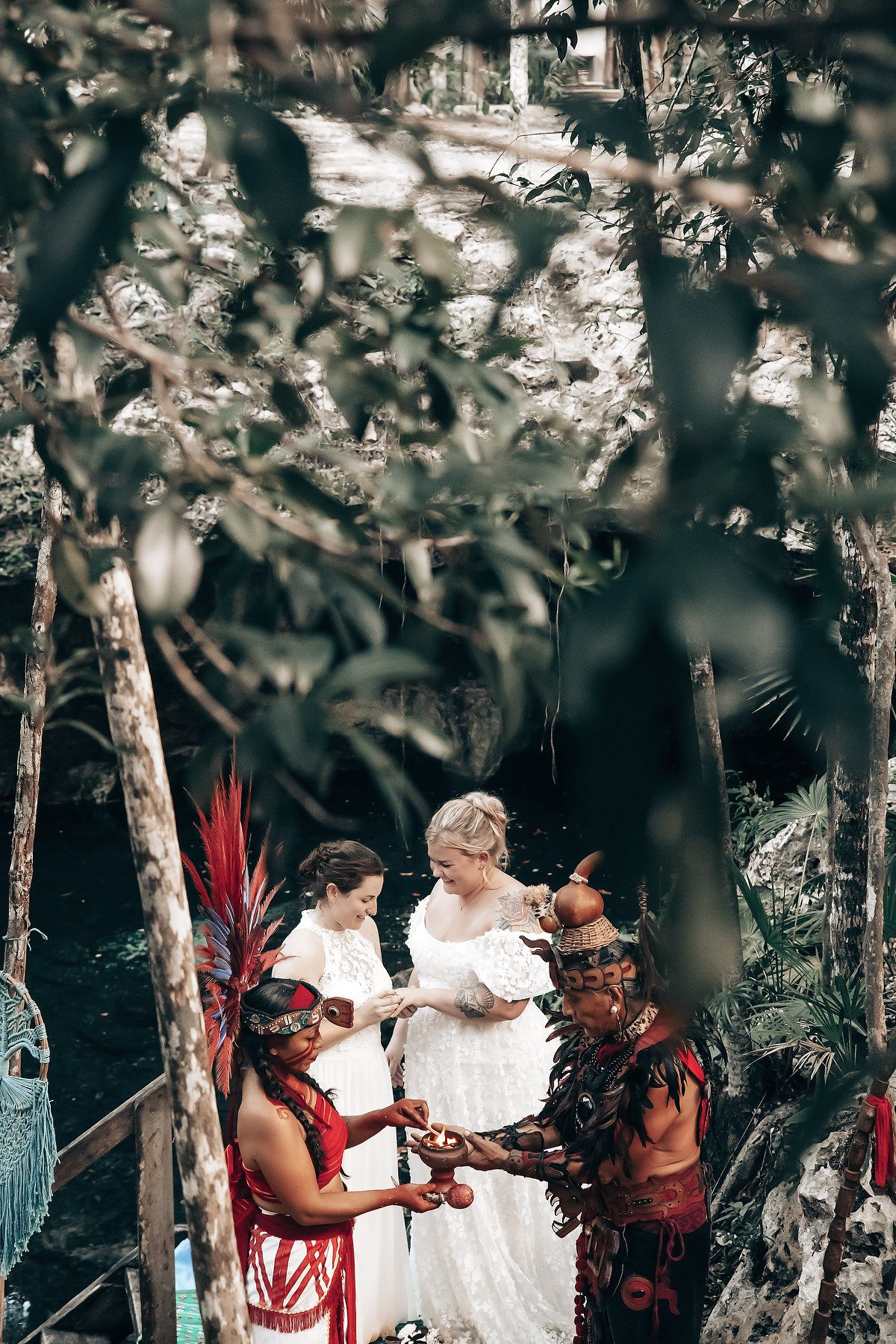 The image shows a bride and groom standing in a forest setting, with a person wearing a colorful, ornate costume in the foreground.