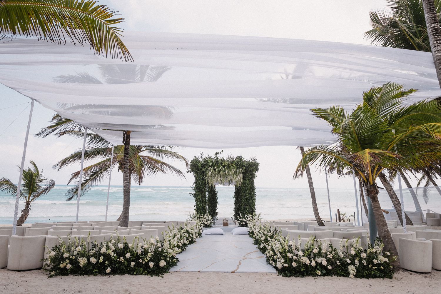 Elegant beach wedding ceremony setup with white floral aisle, greenery arch, palm trees, and draped white canopy by the ocean.