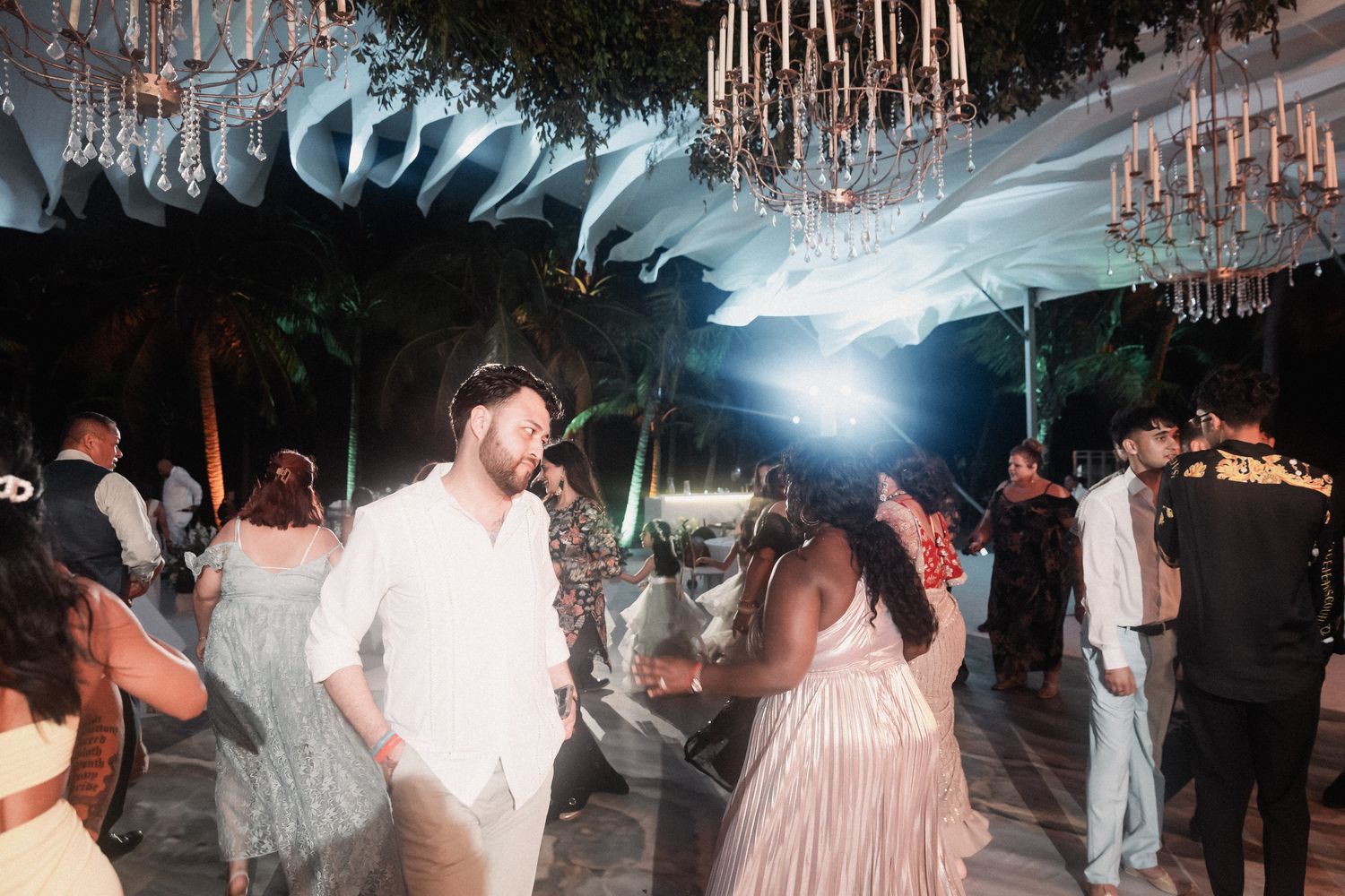 Guests dance energetically under chandelier canopy at lively outdoor tropical wedding reception at night.