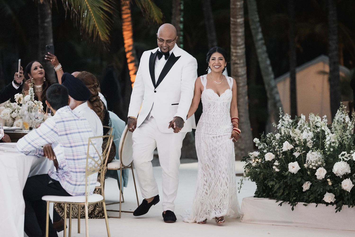 Bride and groom in white wedding attire entering outdoor tropical reception venue.