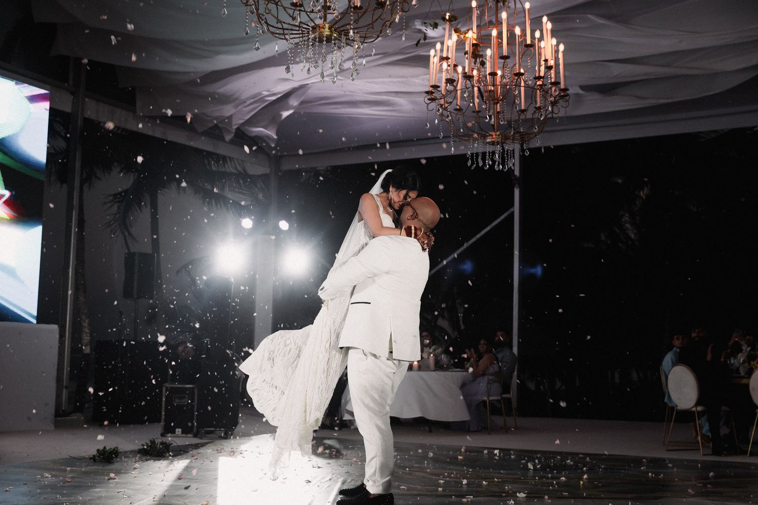 Bride and groom share romantic first dance lift under chandelier with confetti falling at elegant wedding reception.