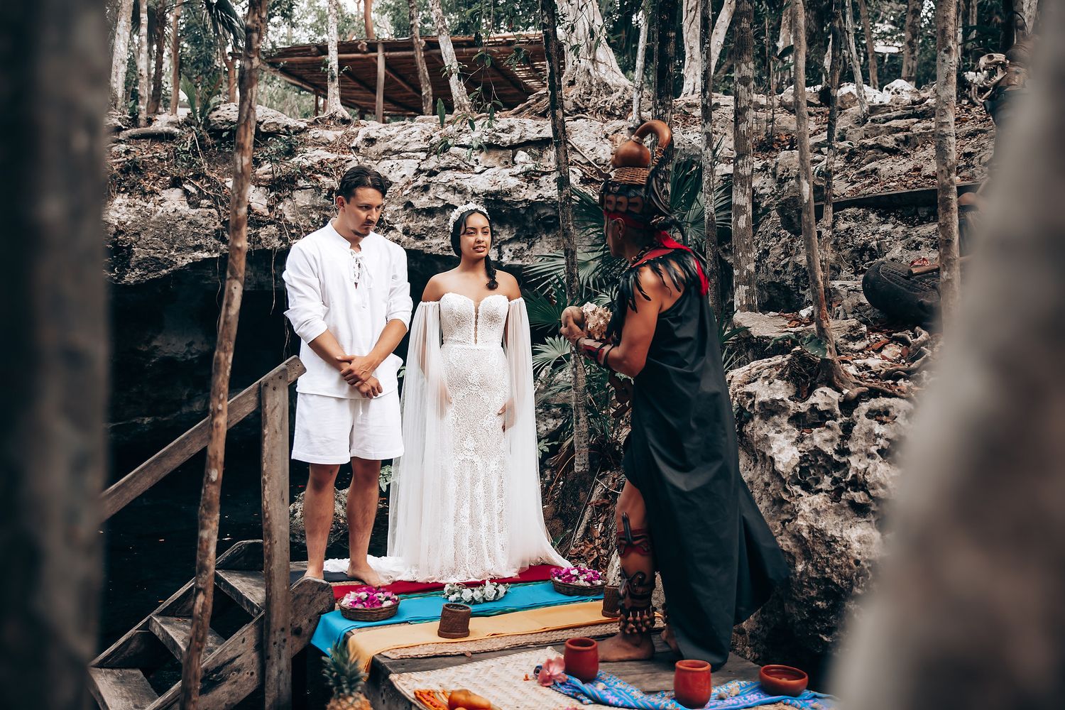 Couple participates in a traditional Hawaiian wedding ceremony on a rocky beach surrounded by tropical foliage.