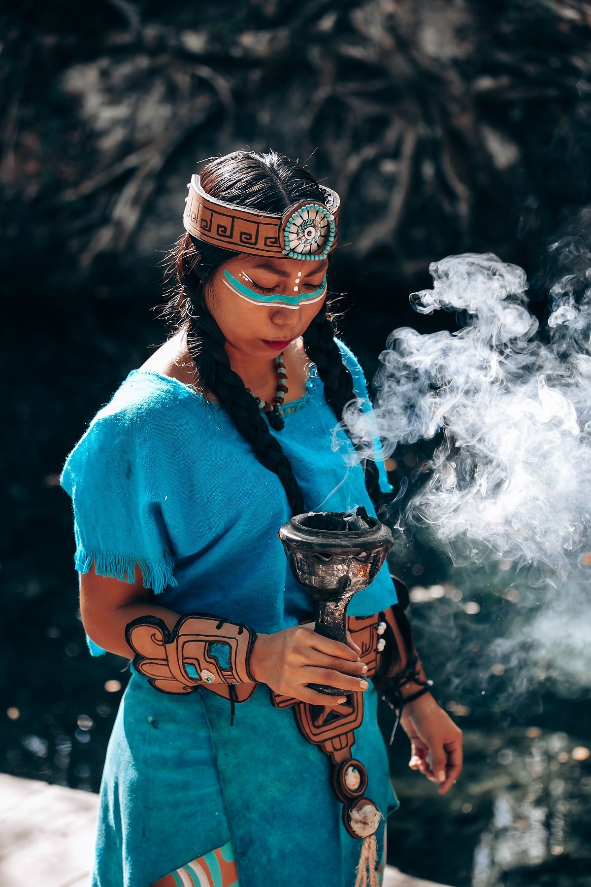 A person in a turquoise shirt and headband performs a ceremonial ritual with smoke and a traditional vessel.