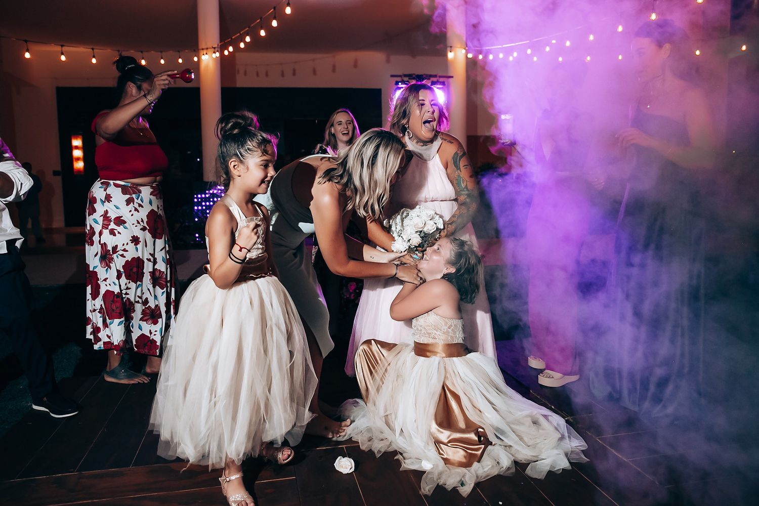 Wedding party dancing on a purple-lit dance floor with fog effects and string lights overhead.