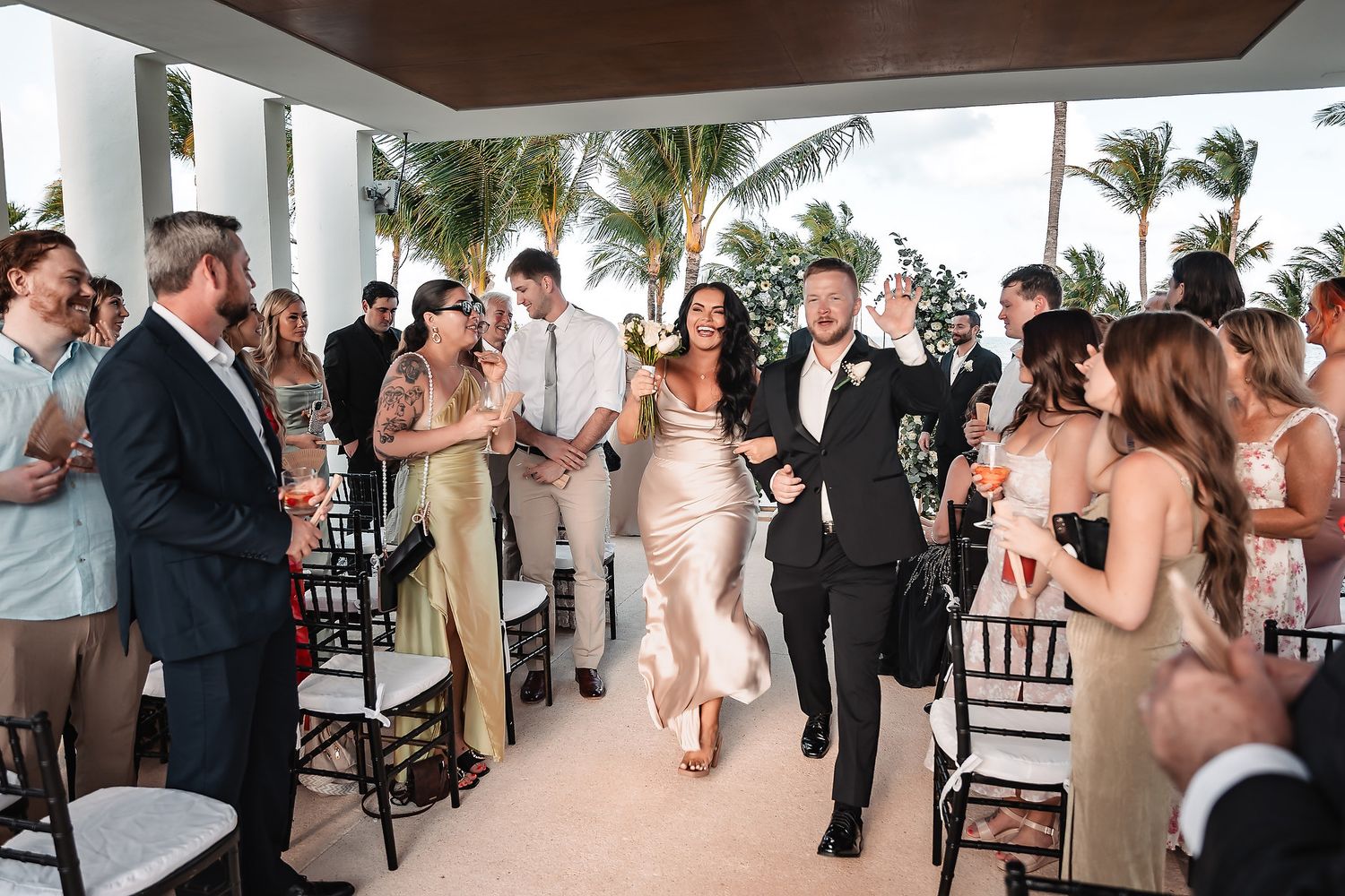 Bride and groom joyfully walk down the aisle at an outdoor tropical wedding reception with palm trees.