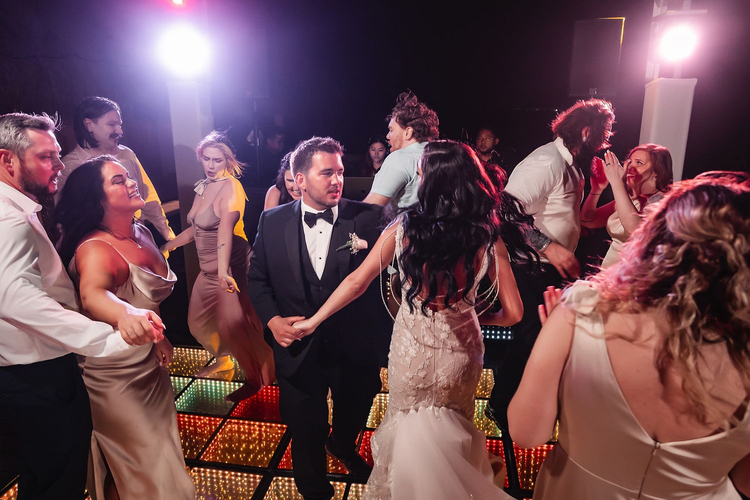 Bride and groom share a dance surrounded by guests on a colorful lit dance floor at a wedding reception.