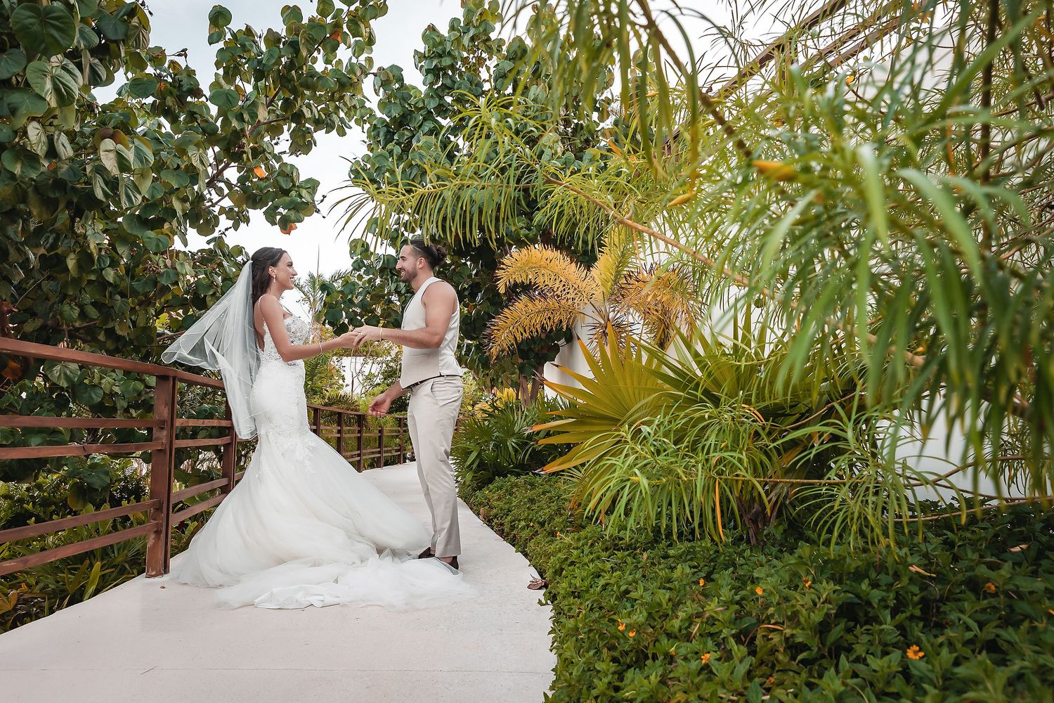 Bride and groom holding hands on a terrace surrounded by lush tropical greenery during an outdoor wedding ceremony.