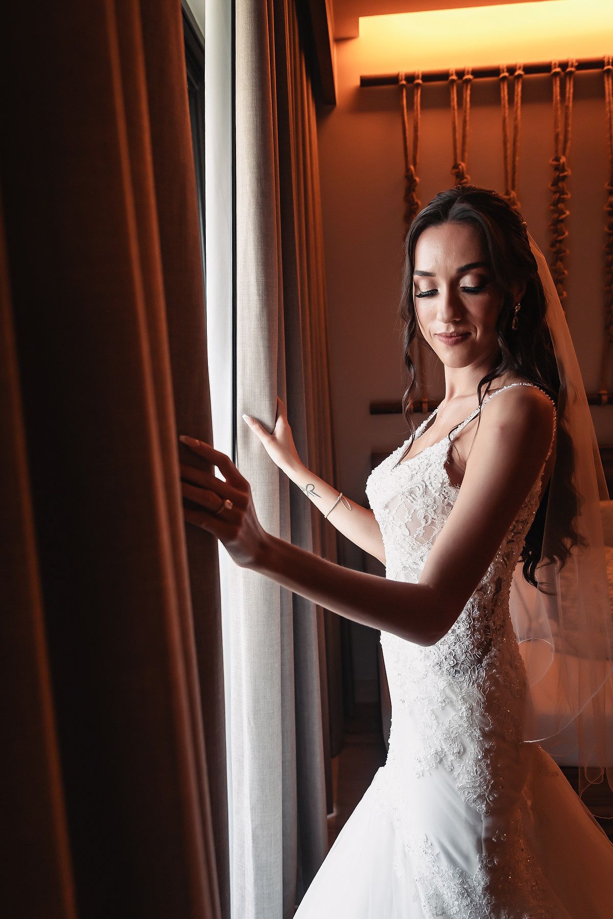 Elegant bride in white lace wedding dress standing by a window, gently touching curtains in soft warm light.