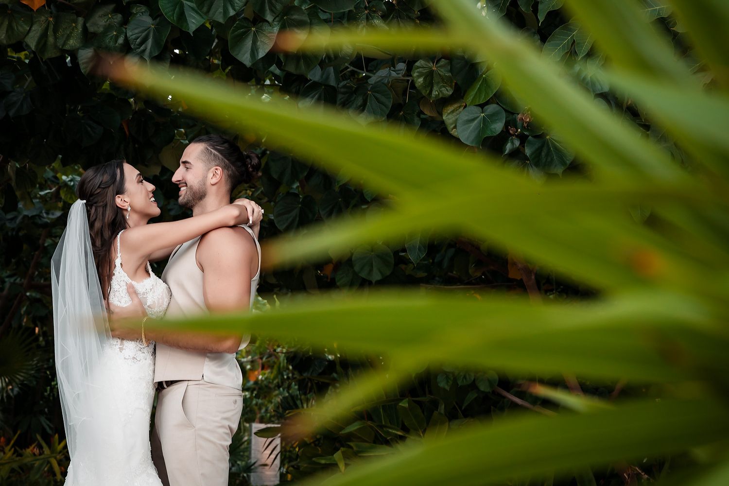 Bride and groom share a romantic embrace among lush tropical foliage at their wedding.