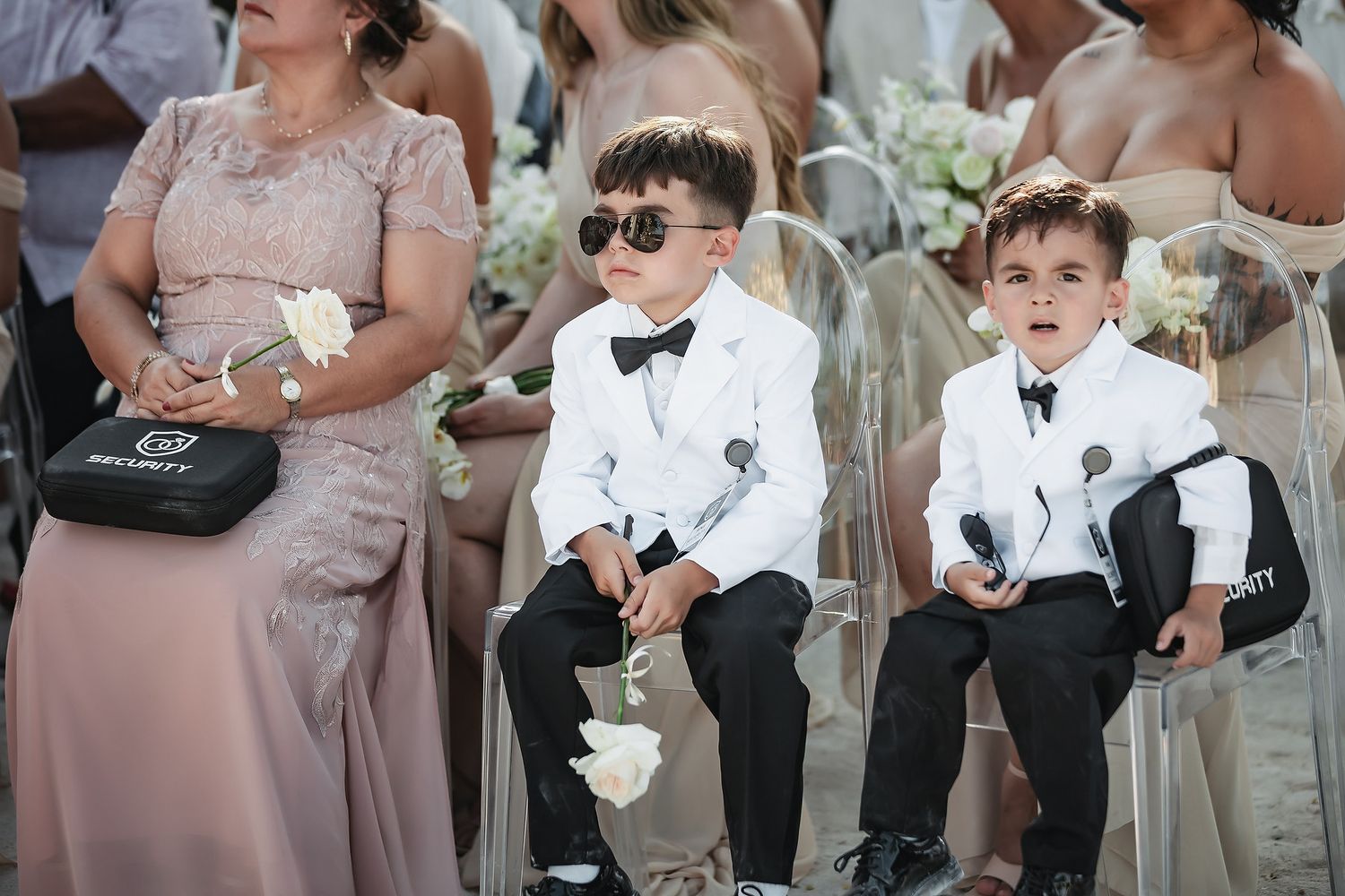 Two young ring bearers in white tuxedos and sunglasses sit at an outdoor wedding ceremony holding small bags.