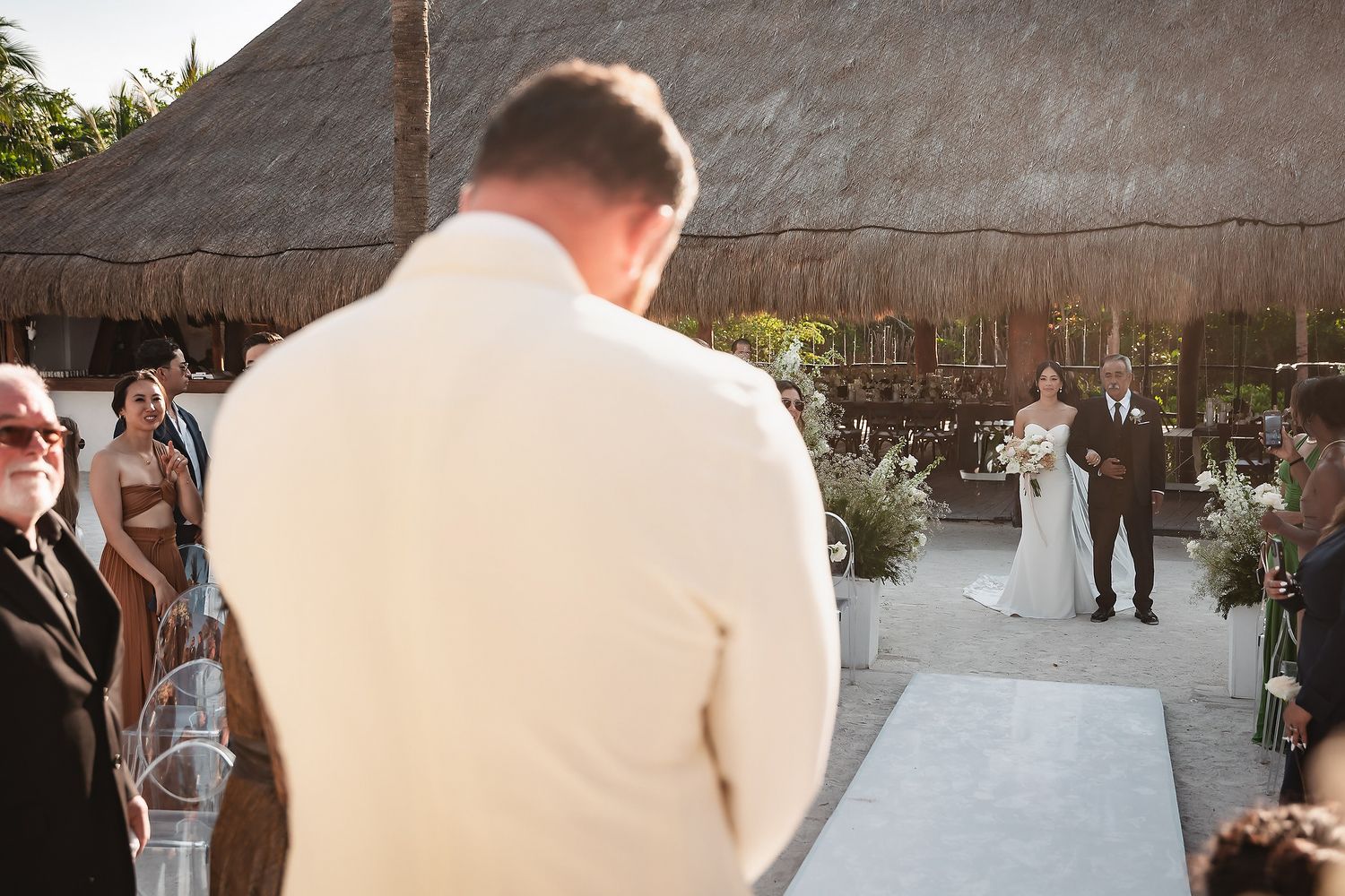Groom stands at altar watching bride walk down aisle at outdoor wedding ceremony with thatched roof venue.