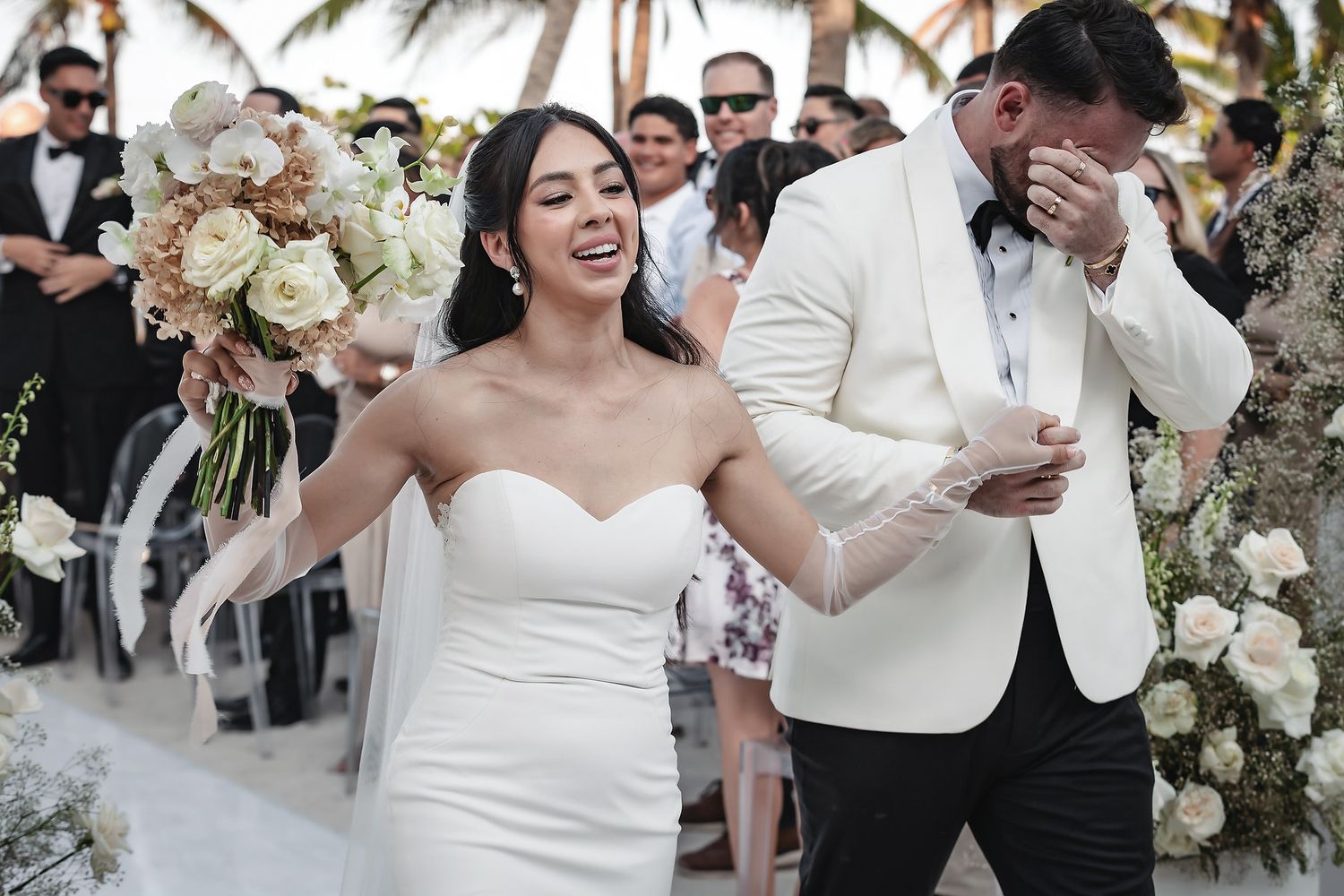 Joyful bride in strapless gown holding floral bouquet with emotional groom in white tuxedo at outdoor beach wedding ceremony.