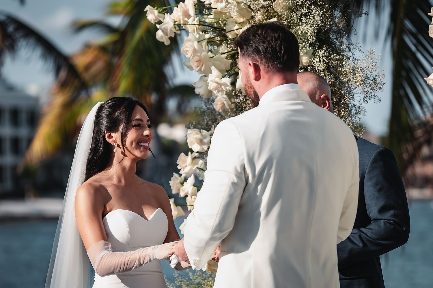 Bride in strapless gown and veil holds hands with groom in white suit at tropical beach wedding ceremony.