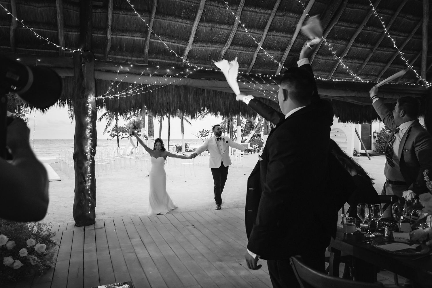 Black and white photo of bride and groom making grand entrance at outdoor beach wedding reception under thatched roof.