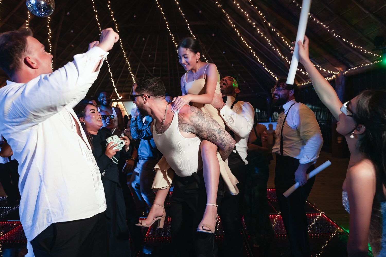 Joyful wedding reception guests lifting bride in the air while dancing under a festive tent with string lights.