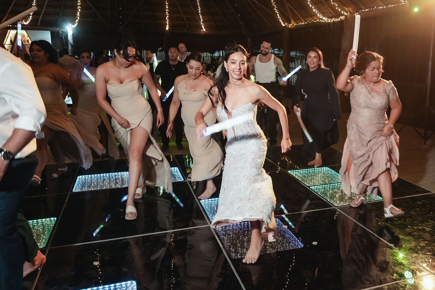 Bride and bridesmaids dancing energetically on a lit dance floor at an outdoor wedding reception.