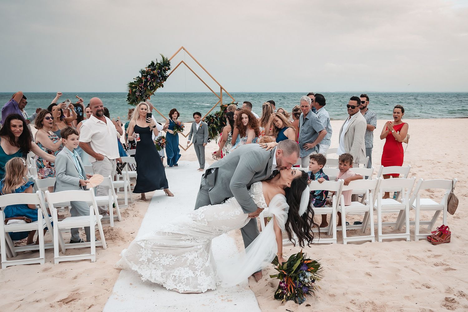 Couple sharing a romantic dip kiss at beach wedding ceremony with guests cheering, geometric arch backdrop, ocean view.