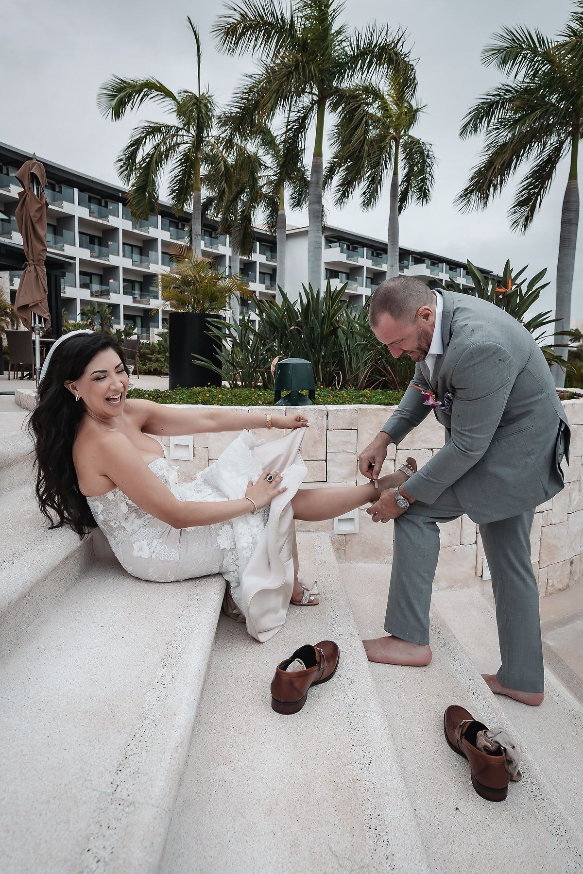 Groom helping bride put on sandals on resort steps, palm trees and hotel in background, tropical wedding.