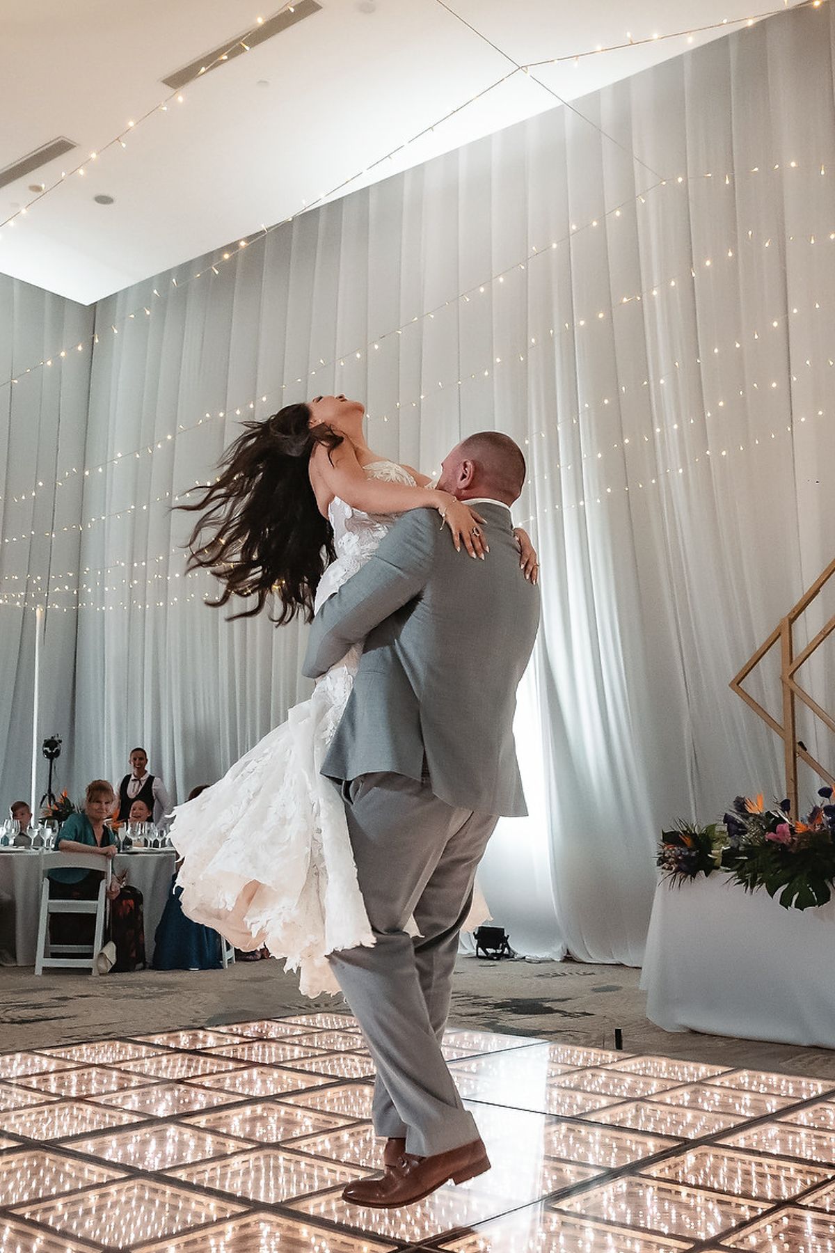 Groom lifts bride during first dance at elegant wedding reception with twinkling fairy lights backdrop.
