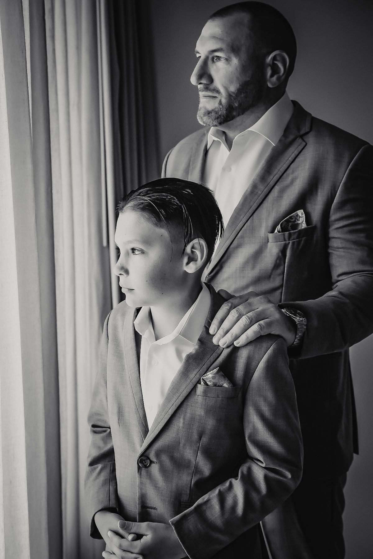 Black and white photo of a father and son in matching suits standing by a window, sharing a quiet moment.