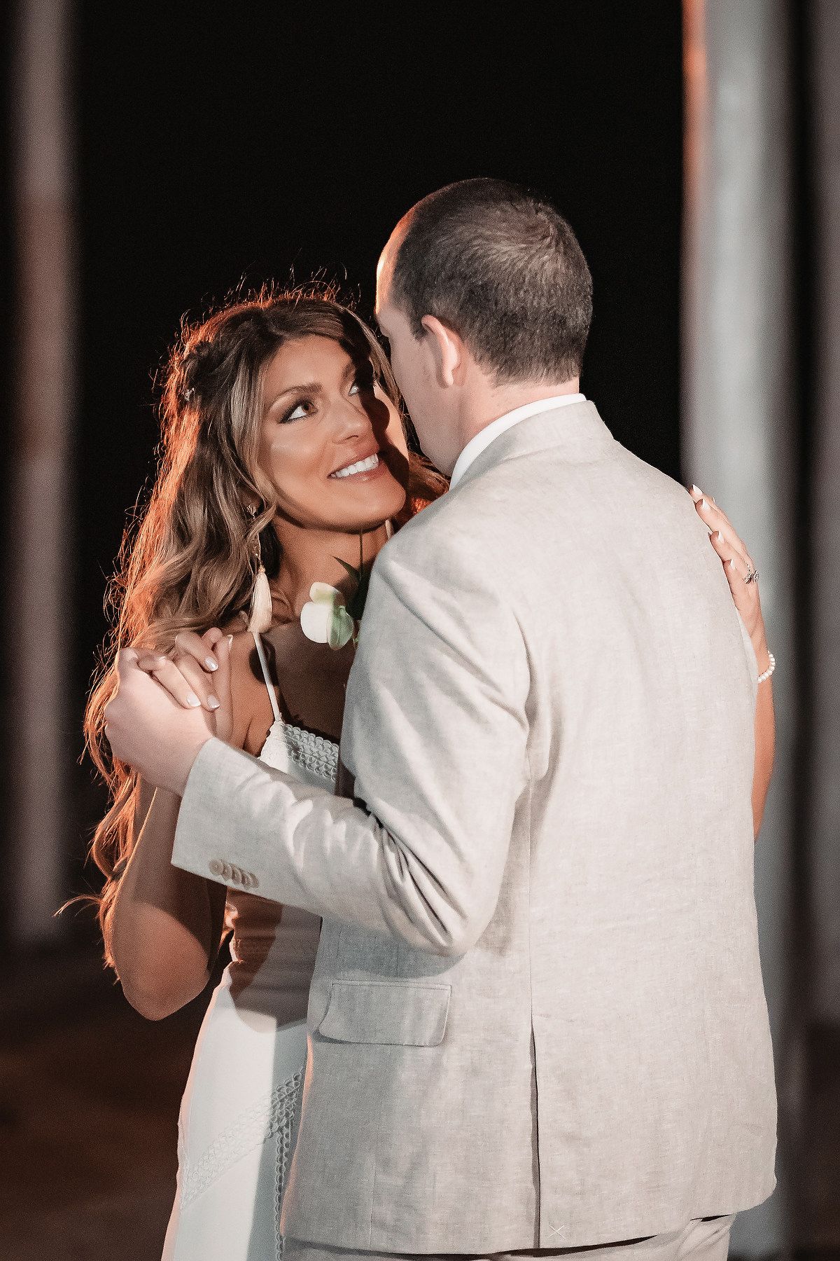 Bride smiling joyfully as groom whispers in her ear during their first dance at a romantic evening wedding reception.