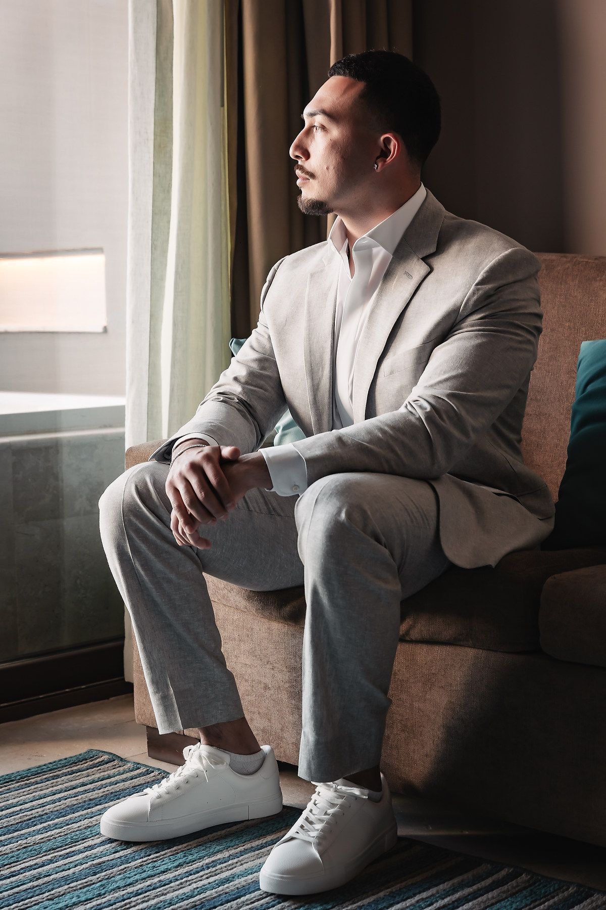 Man in grey suit sitting in armchair gazing out window, wearing white sneakers in elegant hotel room.