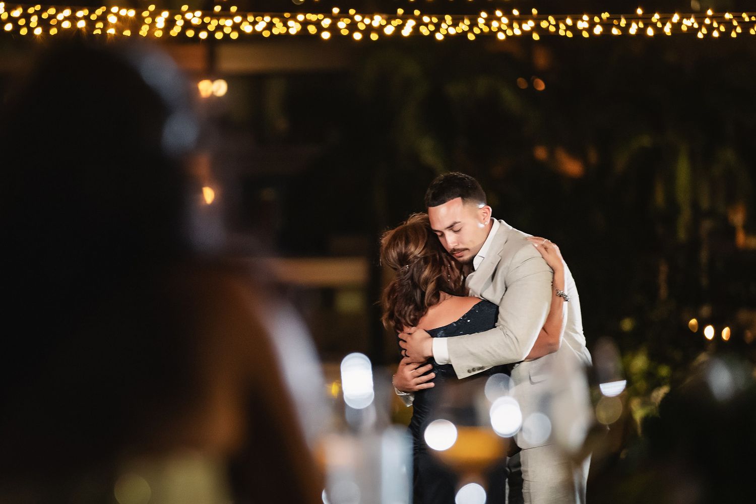 Couple sharing a romantic first dance embrace under warm string lights at an outdoor evening wedding reception.