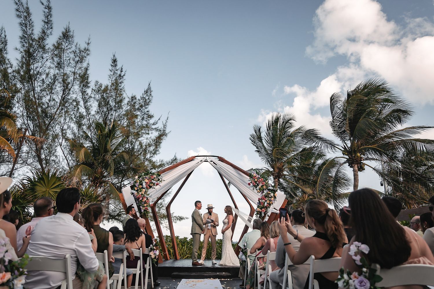 Outdoor beach wedding ceremony with couple at floral arch altar, palm trees, and seated guests under blue sky.