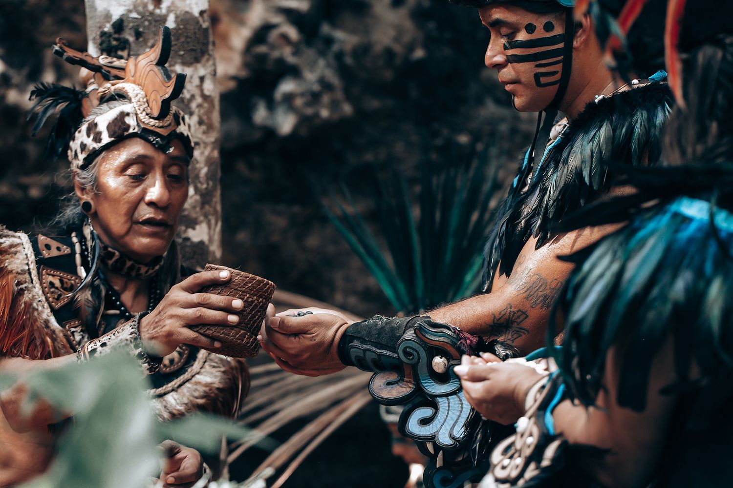 Elder and younger performer in traditional Mayan ceremonial dress share a meaningful exchange in a jungle setting.