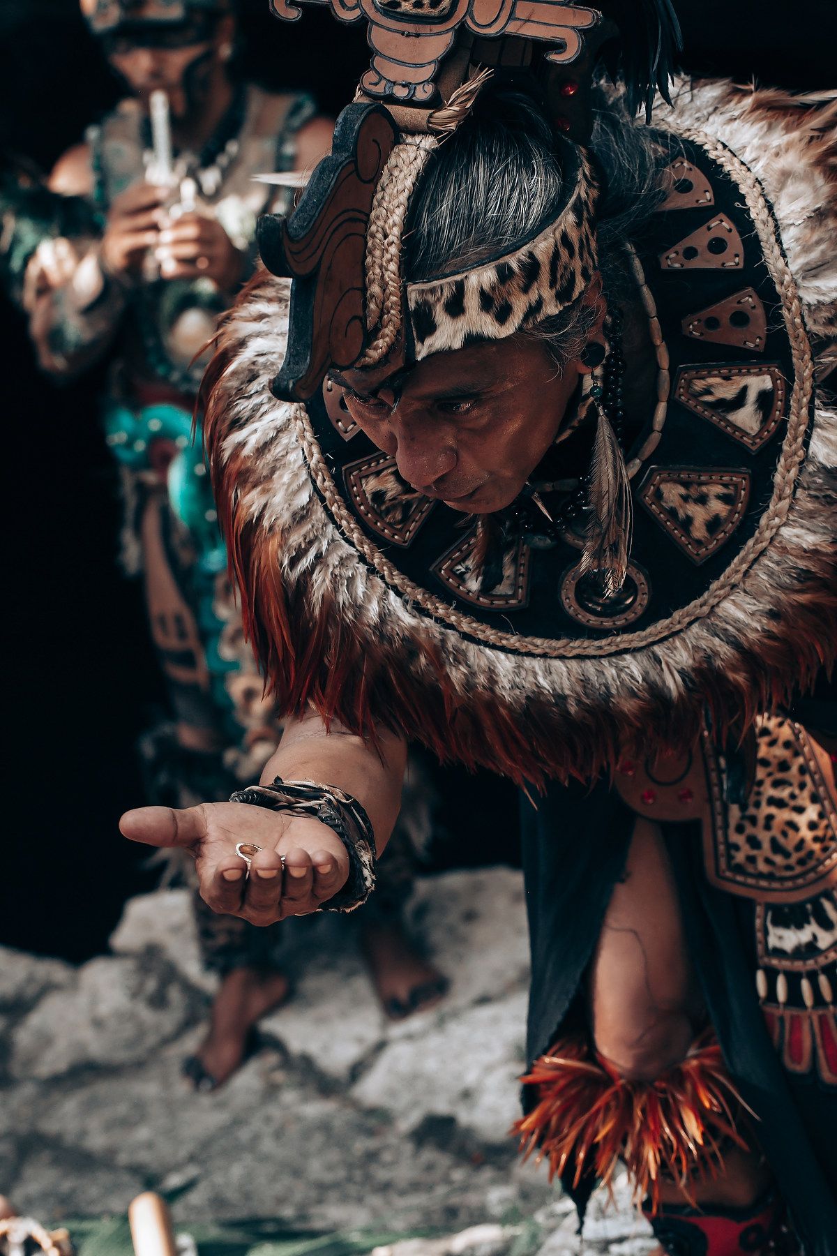 Elder in traditional Mayan feathered headdress and leopard print costume extending hand during a ceremony.
