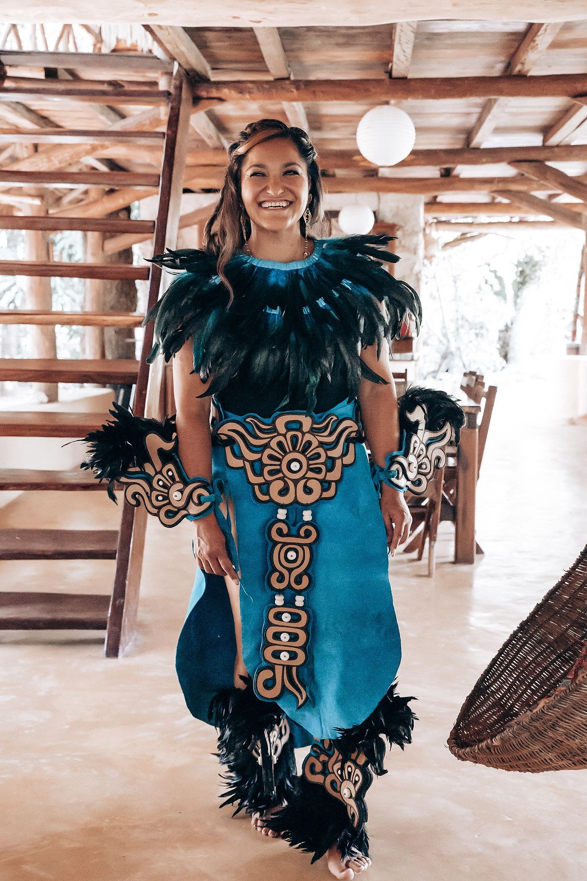 Smiling woman in ornate blue Mayan costume with black feathers and gold embroidery indoors.