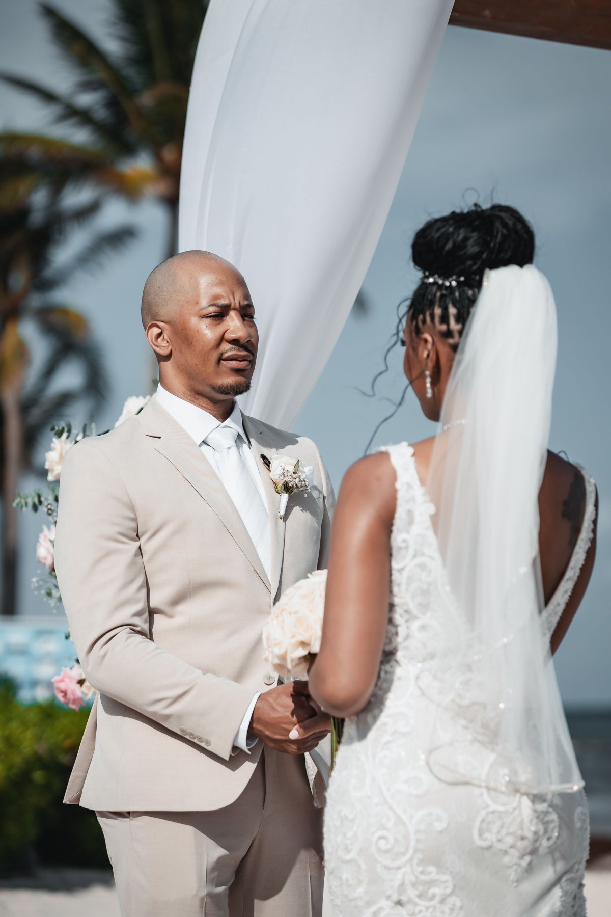 Groom in beige suit holds bride's hands during outdoor beach wedding ceremony under white draped arch.