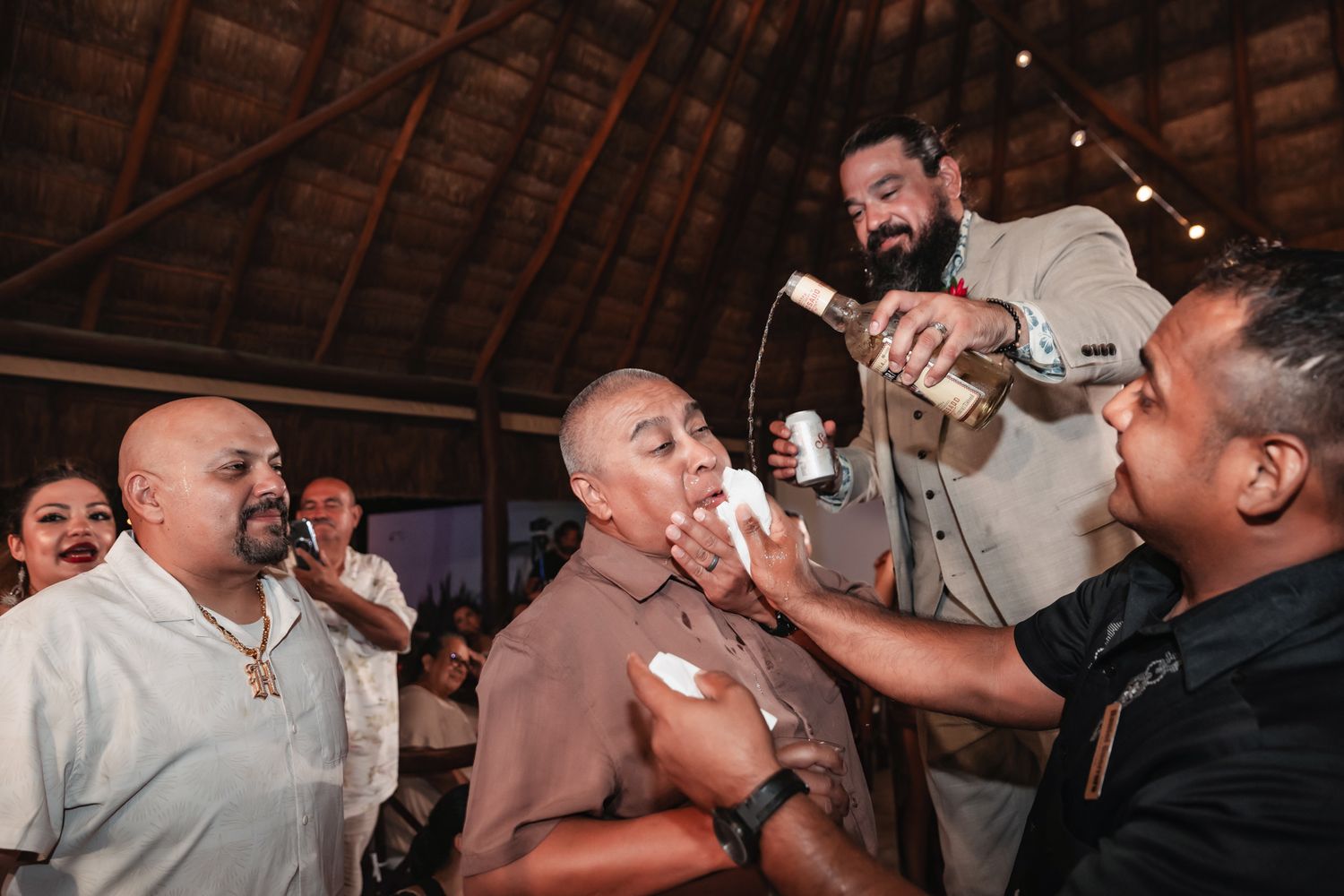 Group of people celebrating indoors under thatched roof, man pouring drink into another person's mouth at party.