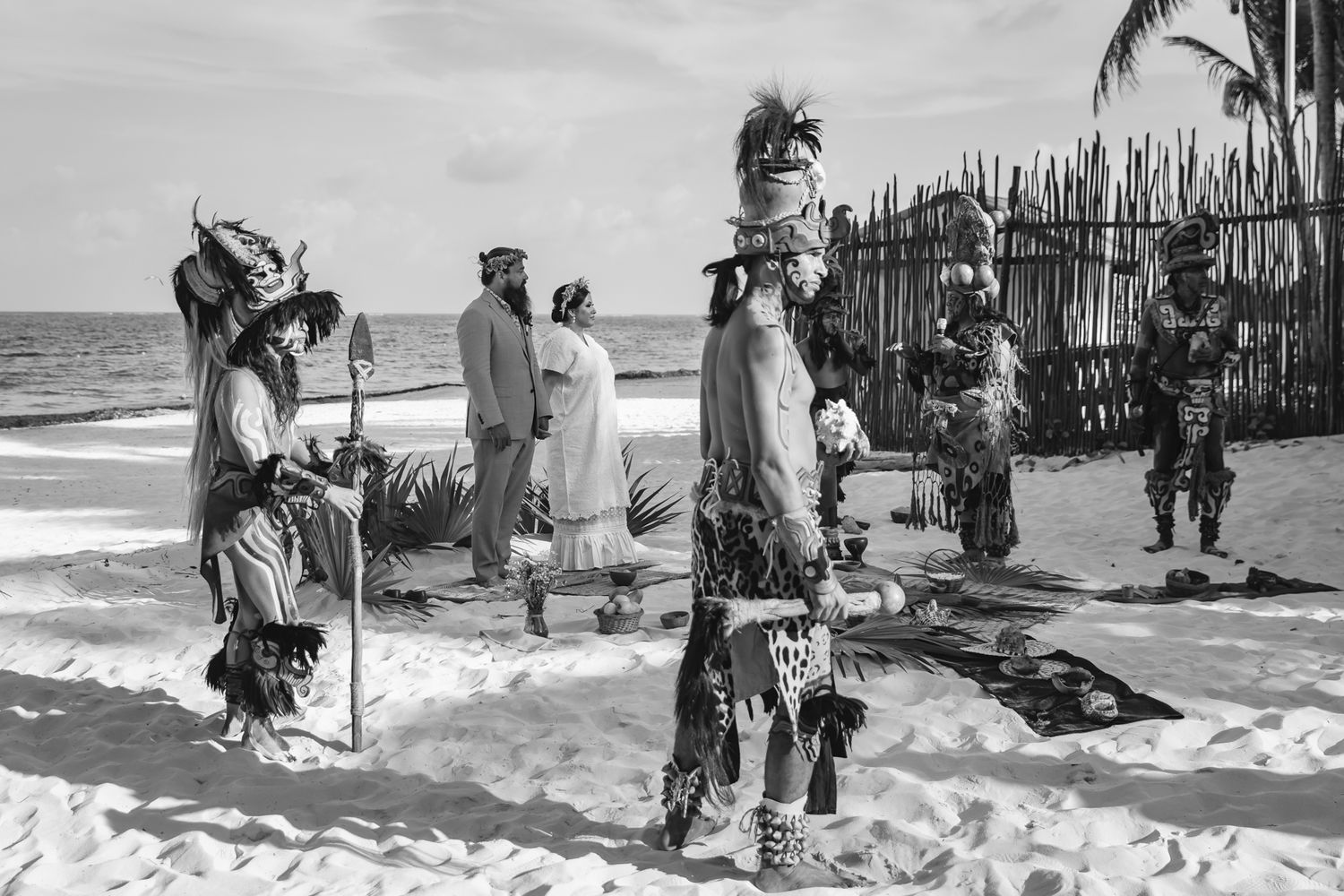Black and white photo of a traditional Mayan ceremony on a beach with costumed performers and ocean backdrop.