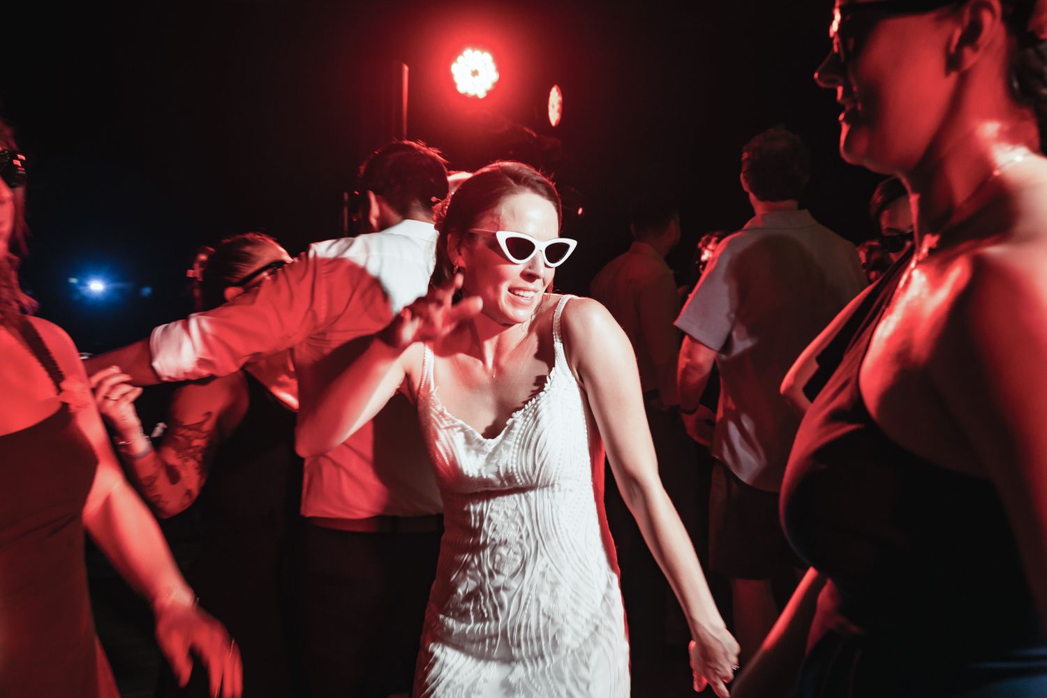 Woman in white dress and cat-eye sunglasses dancing joyfully at a vibrant, red-lit party with guests around her.