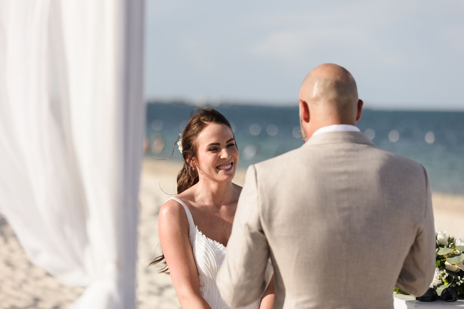 Bride smiling at groom during outdoor beach wedding ceremony with white draping.