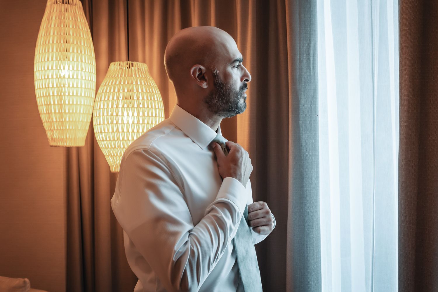 Bald groom adjusting tie by window in hotel room before wedding ceremony.