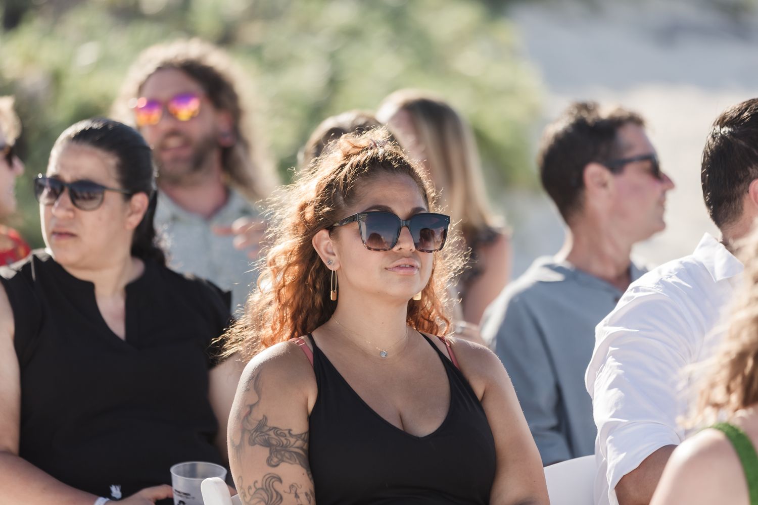 Woman with red curly hair and sunglasses at an outdoor event, tattoos visible, crowd in background.