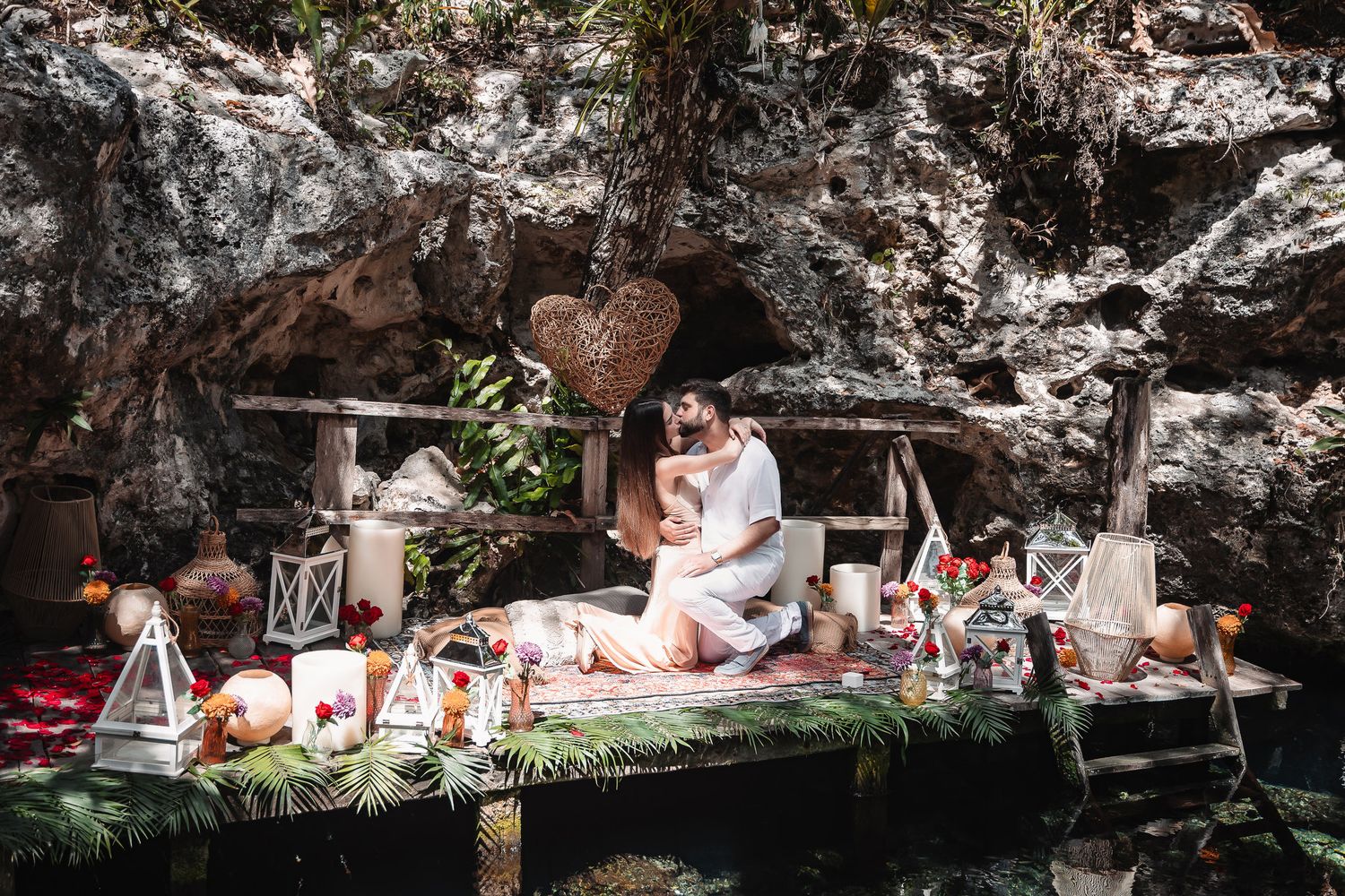 Romantic couple embracing during an intimate outdoor picnic setup with candles, flowers, and wicker heart decor in a natural setting.