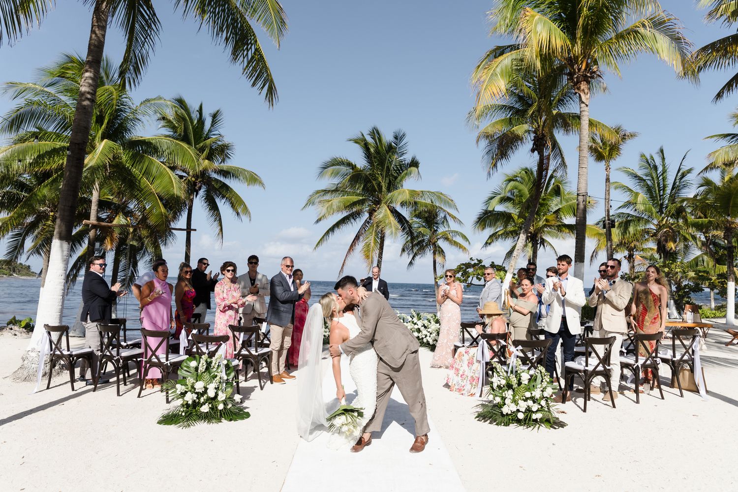 Couple shares first kiss at tropical beach wedding ceremony with palm trees and ocean backdrop.