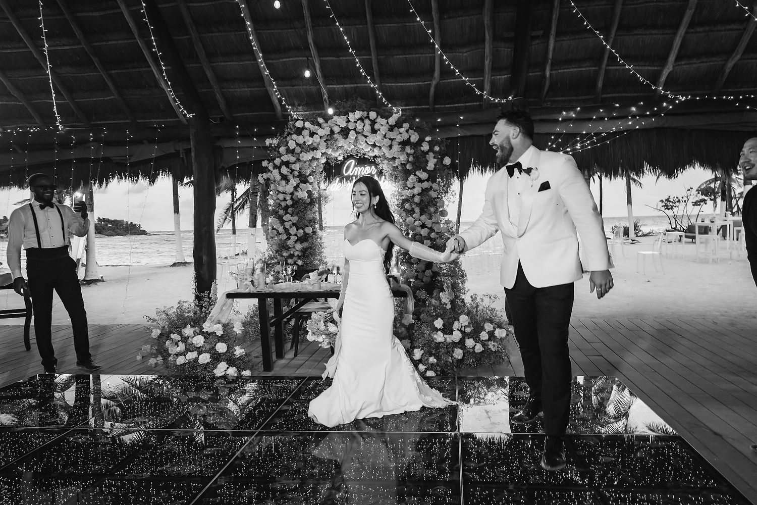 Bride and groom share first dance under twinkling lights and floral arch at elegant outdoor beach wedding reception.