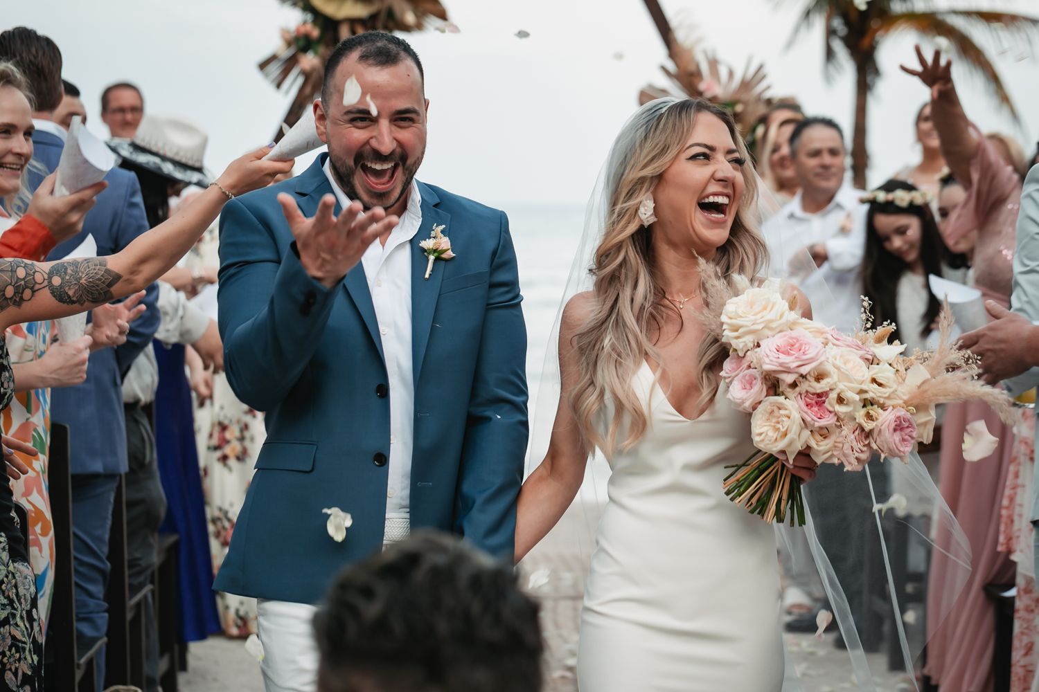 Joyful bride and groom walk down beach wedding aisle as guests celebrate with confetti and cheers.