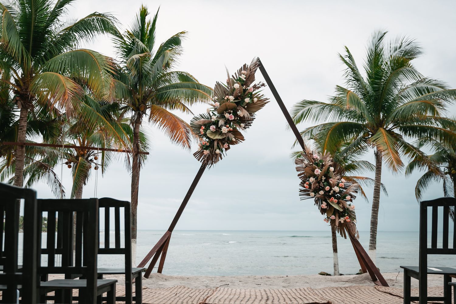 Tropical beach wedding ceremony setup with triangular floral arch, palm trees, and ocean backdrop.