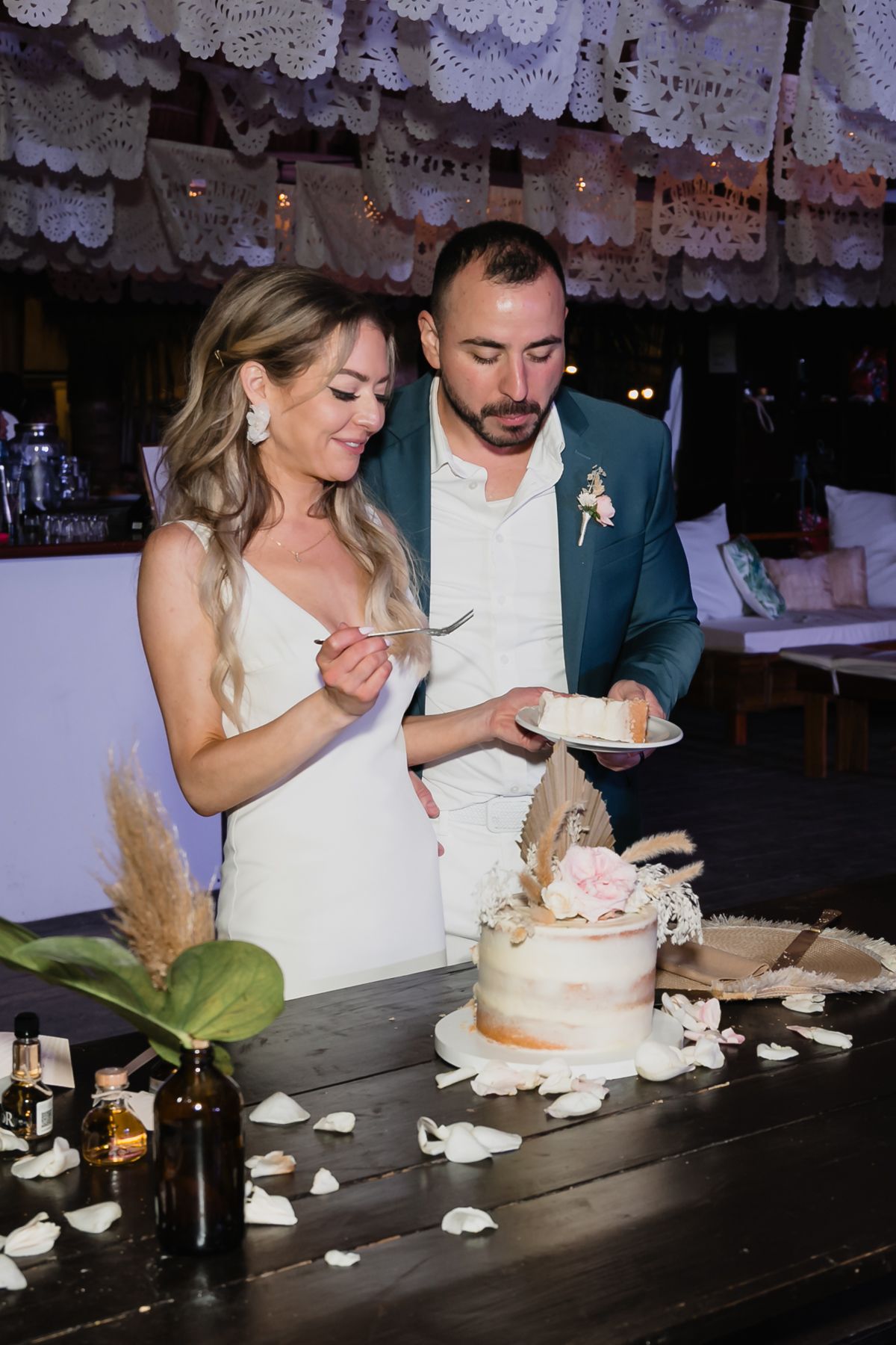 Bride and groom in teal blazer cut naked wedding cake decorated with flowers under papel picado at reception.