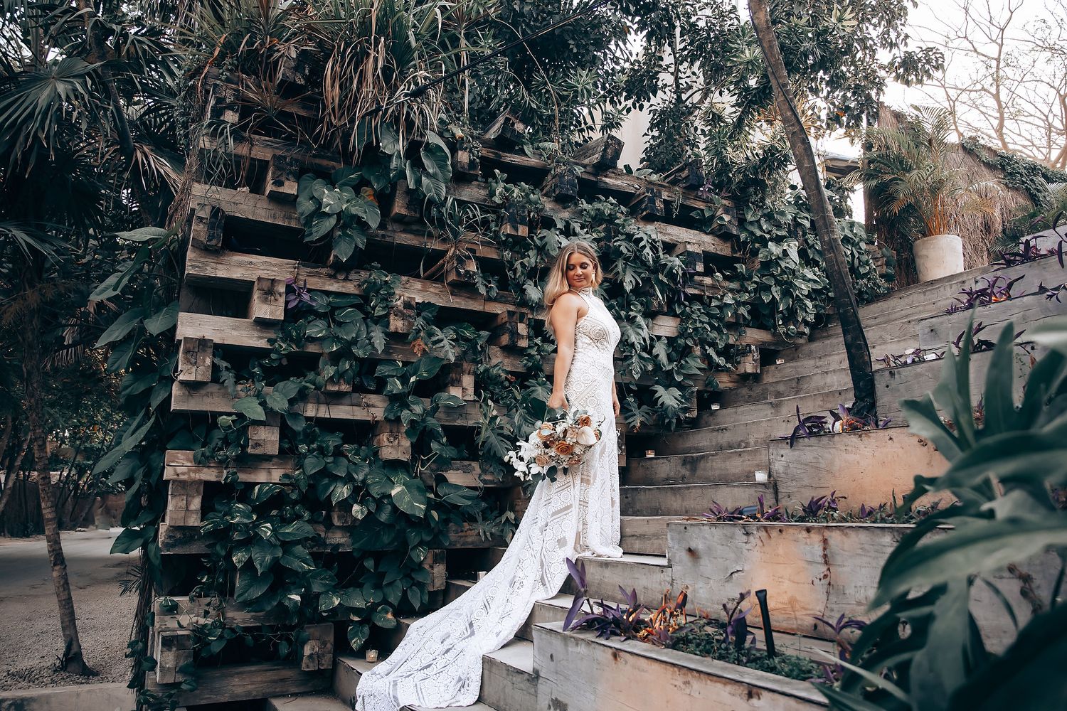 Bride in white dress poses gracefully in a rustic outdoor garden setting with lush greenery and natural lighting.