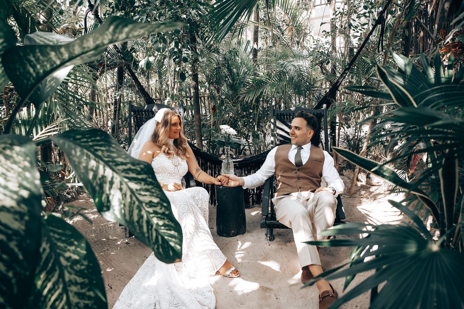 A couple in wedding attire sits together in a lush tropical garden setting with palm fronds surrounding them.