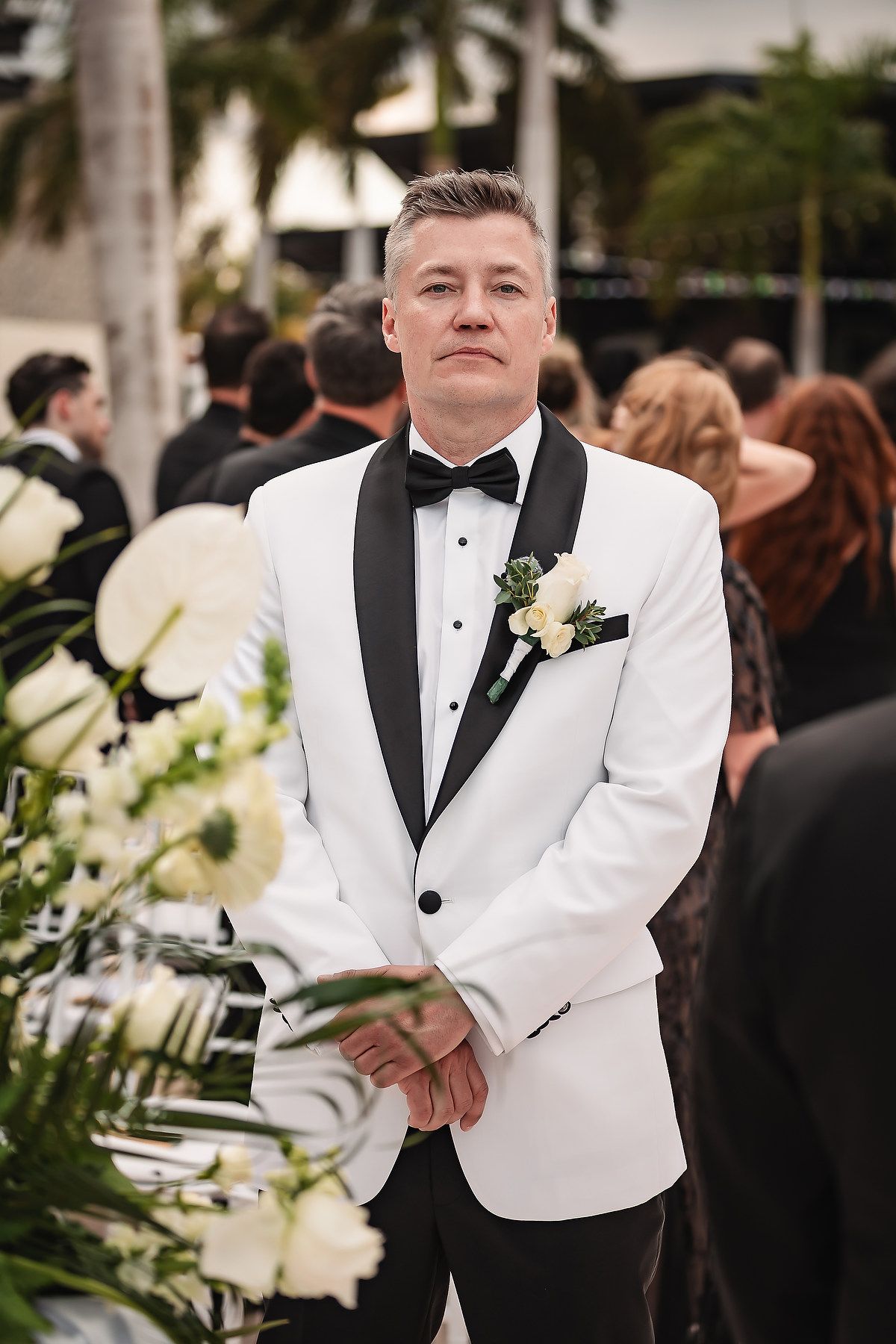 Groom in classic white tuxedo jacket and black bow tie smiles during outdoor wedding ceremony with floral decor.
