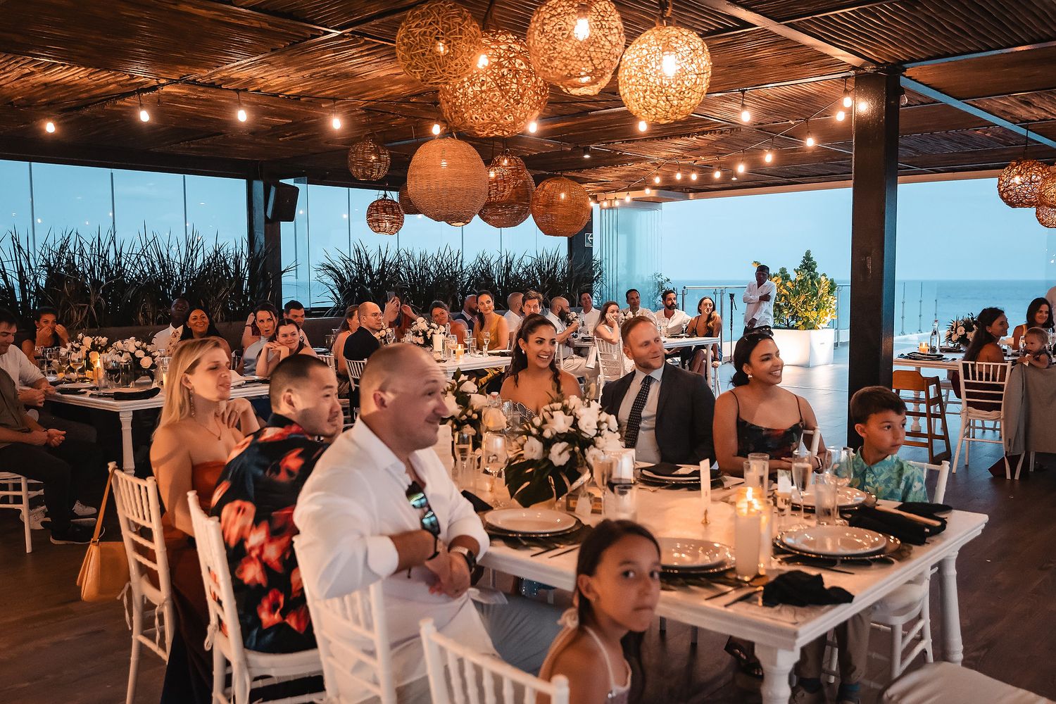 Wedding reception dinner at an oceanfront venue with glowing wicker pendant lights and white table settings.