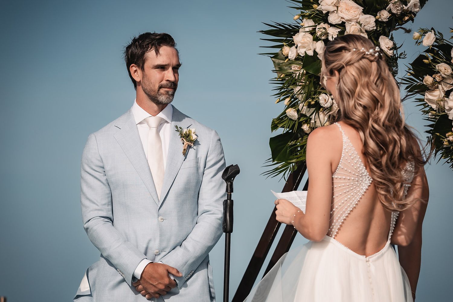 Intimate wedding ceremony moment with couple standing before floral arch against ocean backdrop.