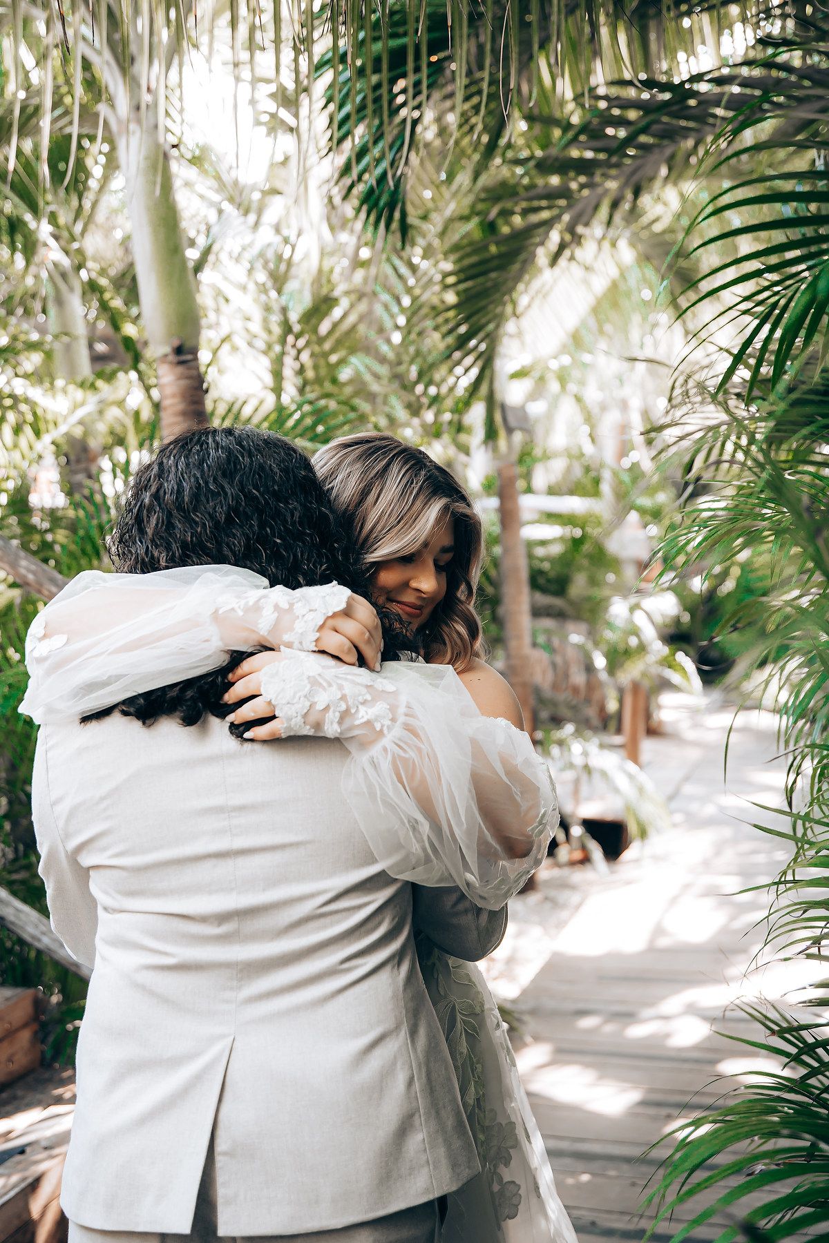Two people share an intimate embrace in a lush tropical garden setting with palm trees in the background.